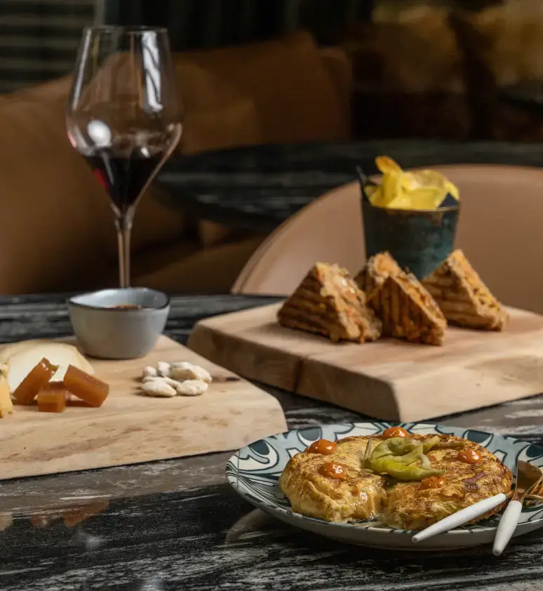 Una taula parada amb una copa de vi negre, un plat de formatges i fruits secs, patates fregides, triangles de sandvitx i un plat de truita espanyola guarnida amb pebrot verd.